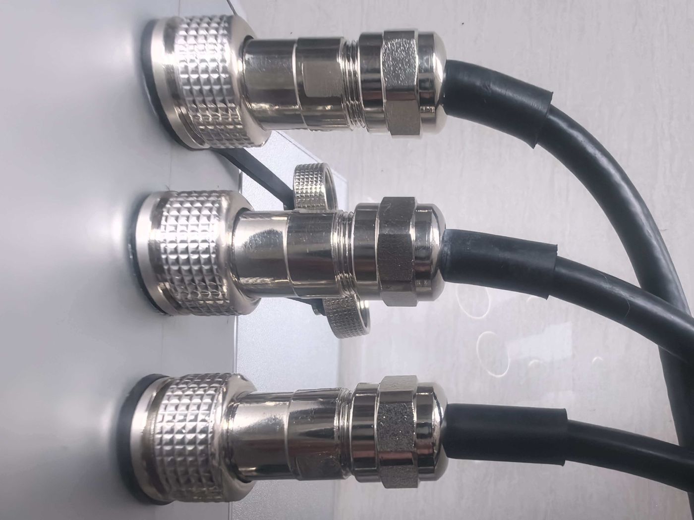 Three metal circular connectors fixed in a row through a white panel, each with a black cable