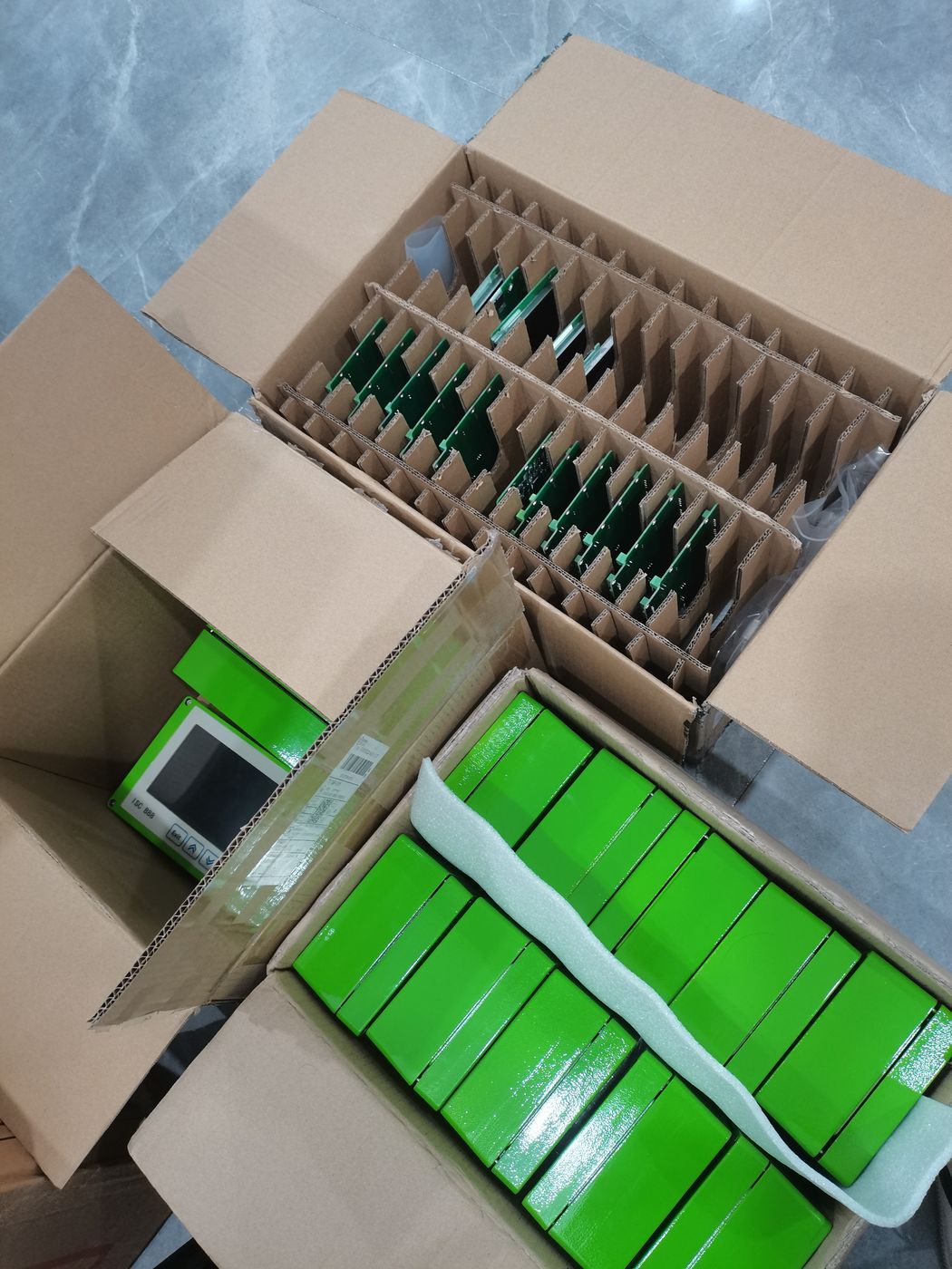 Open carton with small modules standing in dividers and a row of green inner boxes in front