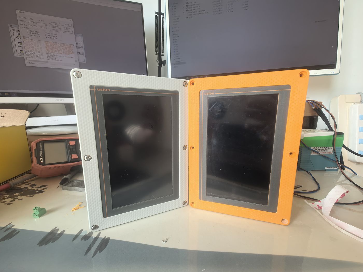 Two terminal face frames standing side by side on a bench, one white and one orange, with monitors behind