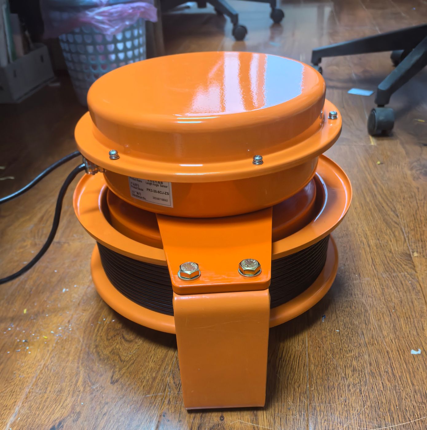 Round orange draw-wire reel with mounting lugs and a nameplate on a wooden floor