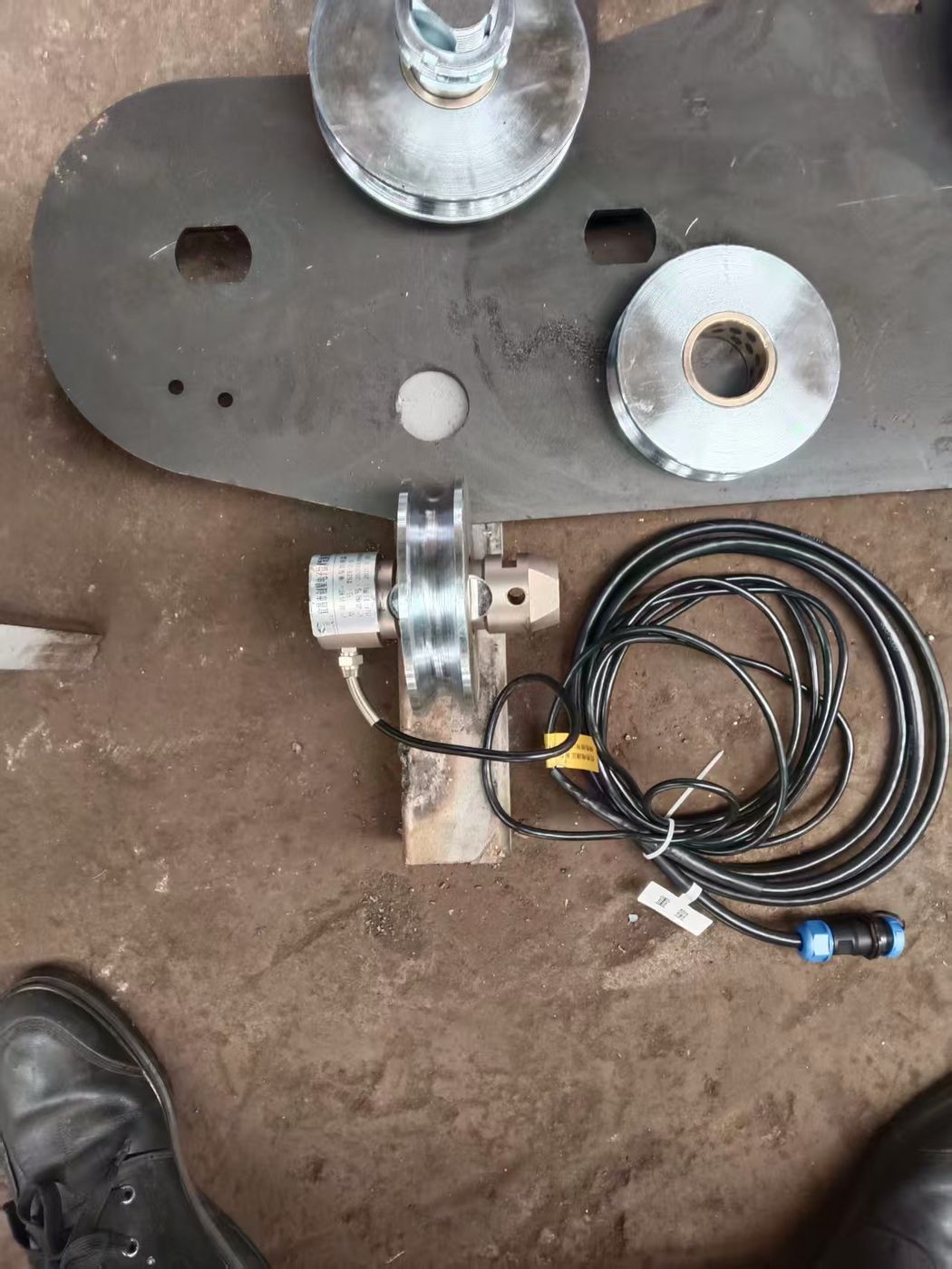 Black plate, two silver bushings and a cell with coiled cable on muddy ground, work boots alongside