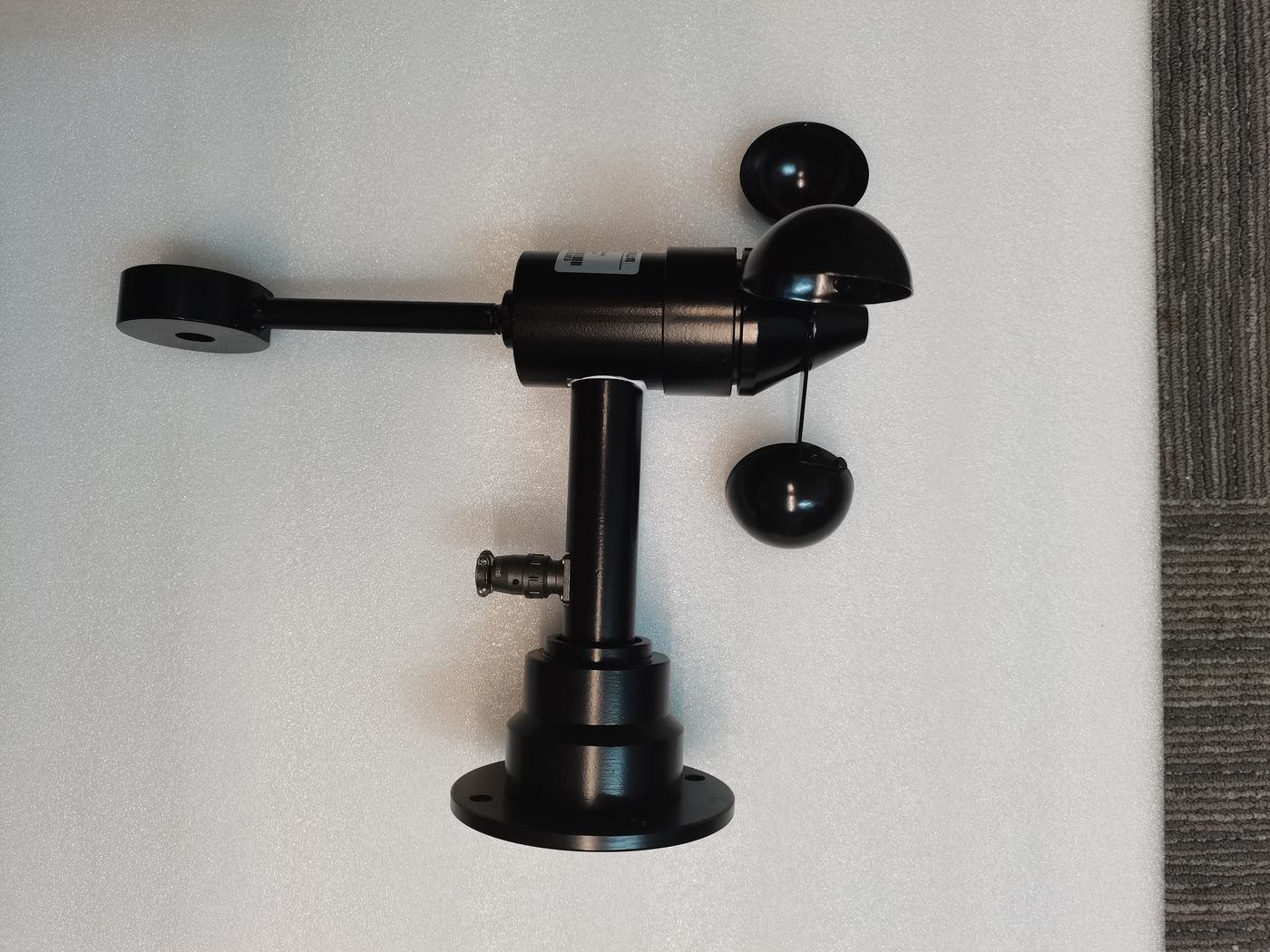 Black cup anemometer with a wind vane and a base flange, lying on white foam