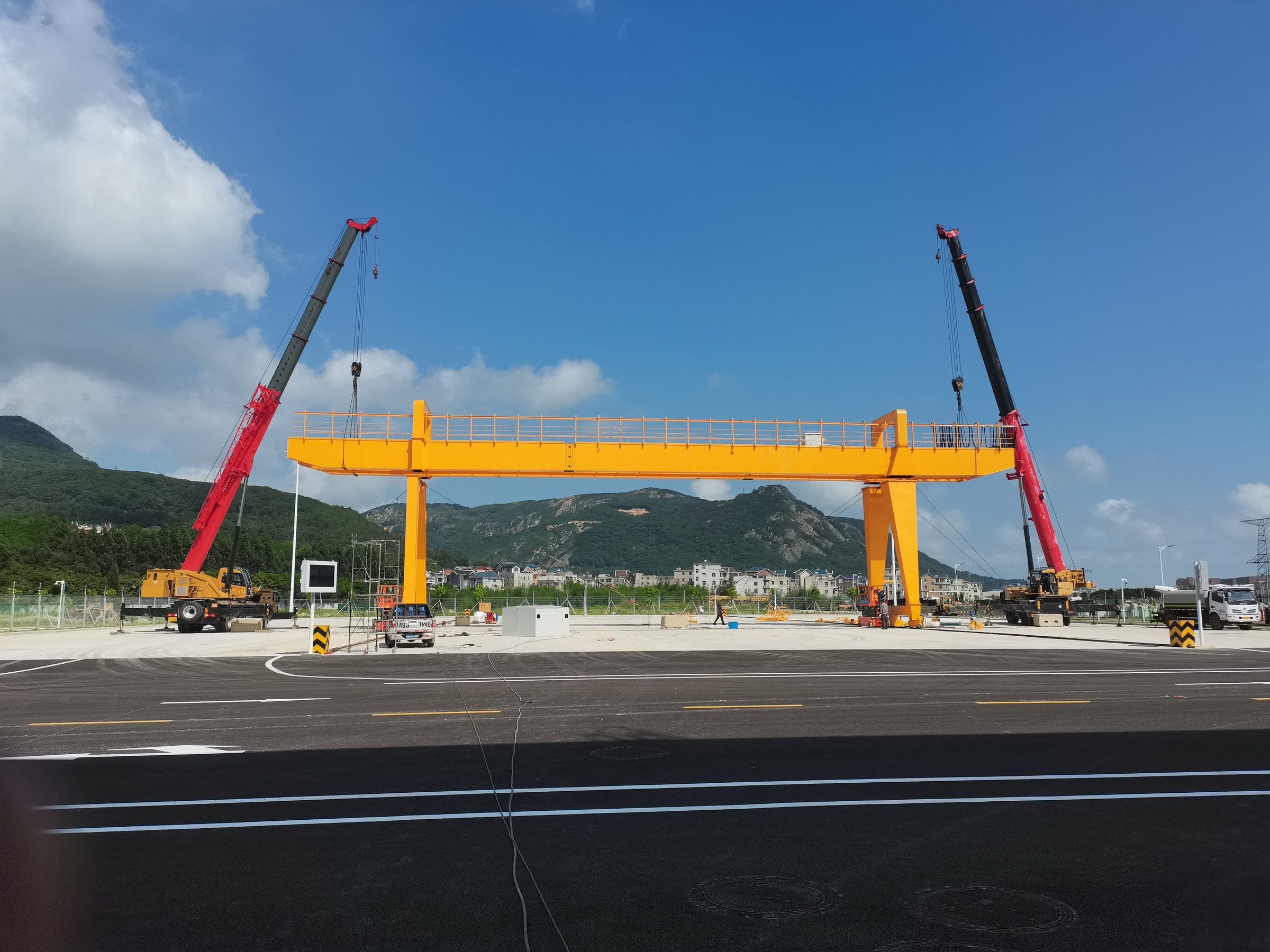 Two mobile cranes erecting an orange gantry leg under a blue sky